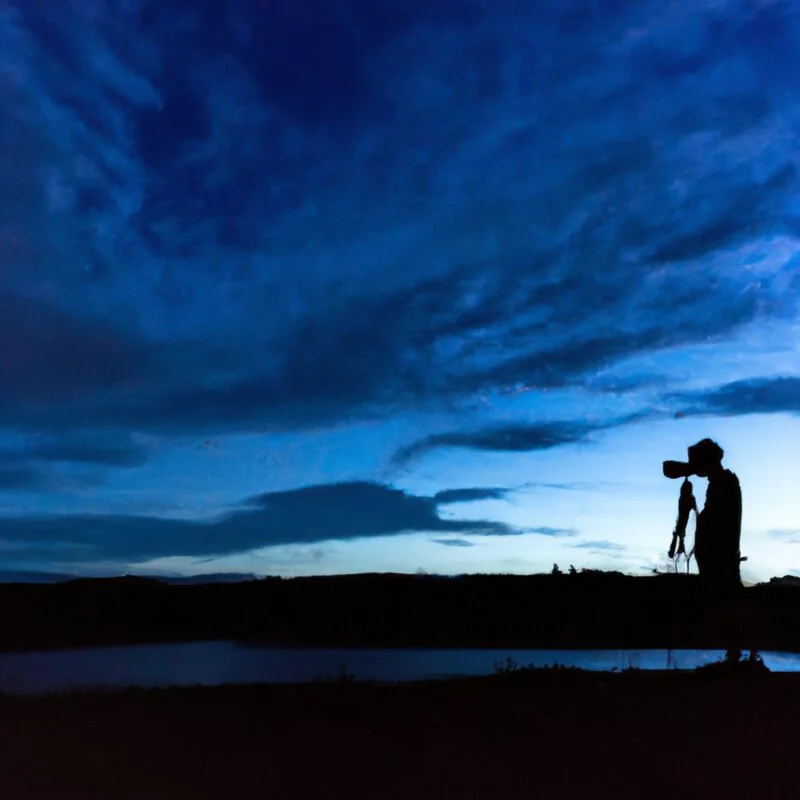 A serene blue hour landscape with a photographer silhouetted against a twilight sky, adjusting a camera on a tripod, with hints of a storm clearing in the background