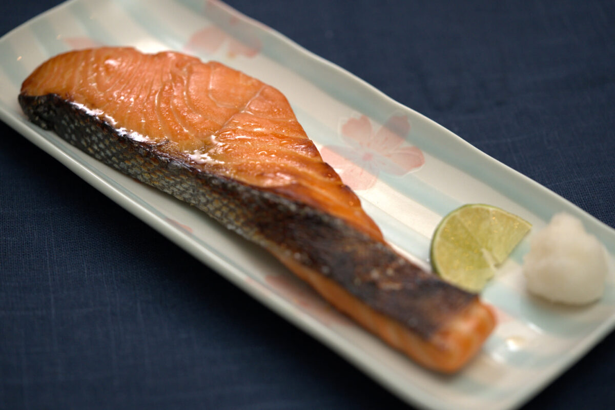 Grilled salmon fillet with crispy skin on a rectangular Japanese ceramic plate with floral motif, garnished with a lime wedge and small white condiment, on a dark blue linen background