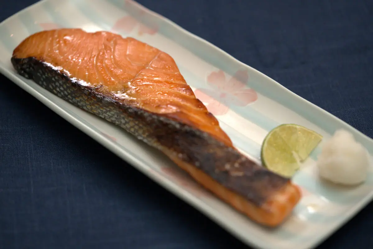 Grilled salmon fillet with crispy skin on a rectangular Japanese ceramic plate with floral motif, garnished with a lime wedge and small white condiment, on a dark blue linen background