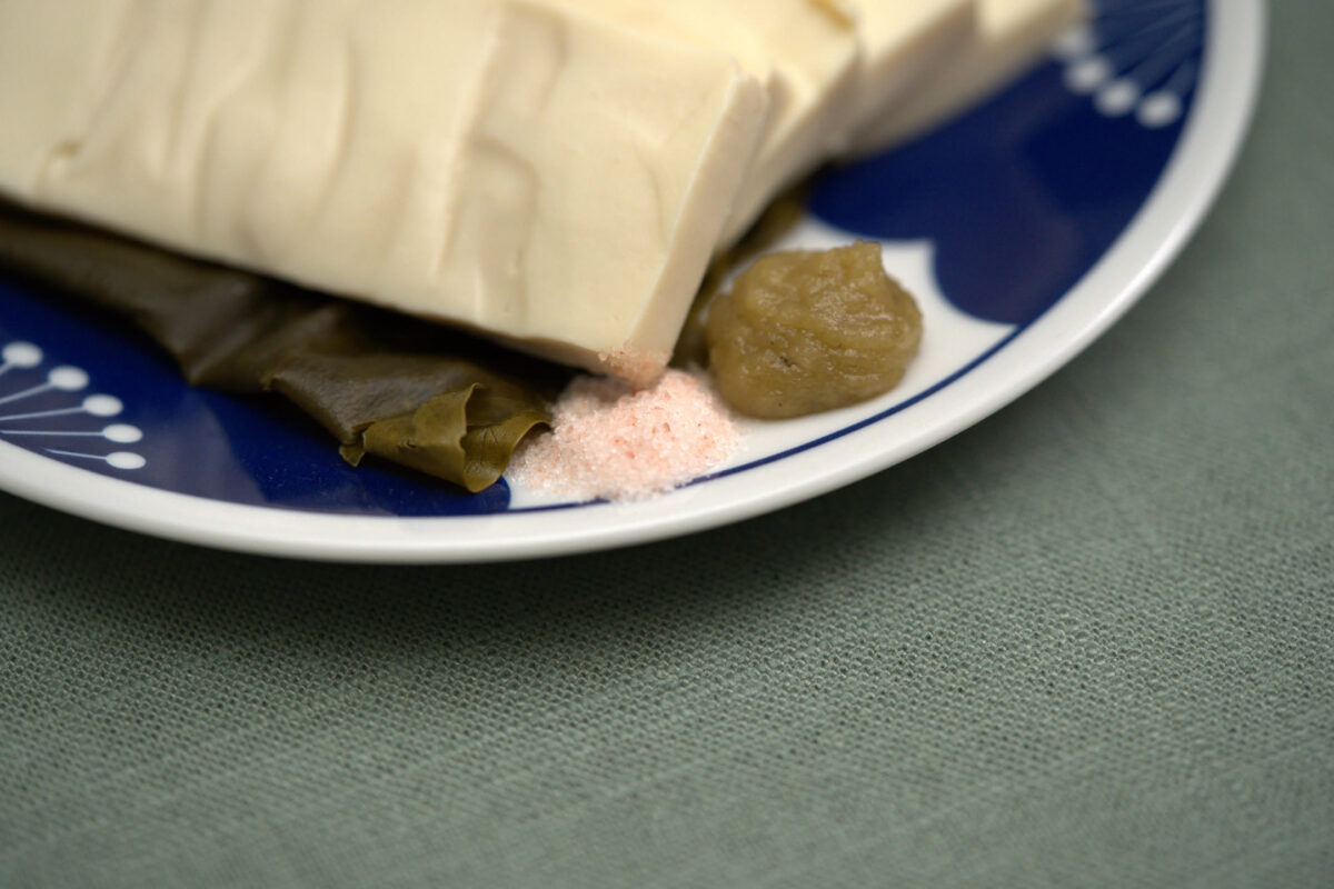 Close-up of a block of silken tofu on a blue and white plate, accompanied by pieces of kombu seaweed, a small mound of pink salt, and a round umeboshi pickled plum