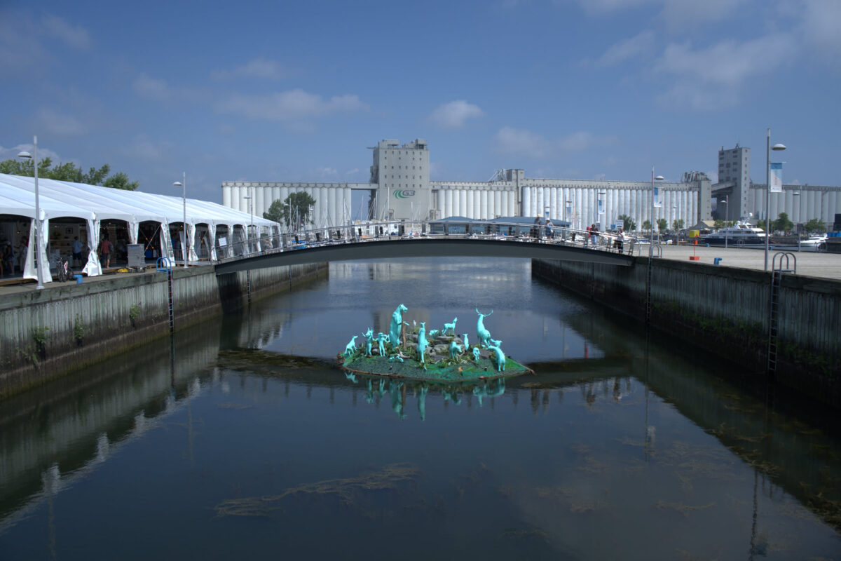Urban canal with a small island featuring turquoise animal sculptures, a pedestrian bridge, industrial grain silos in the background, and event tents along the waterfront on a sunny day