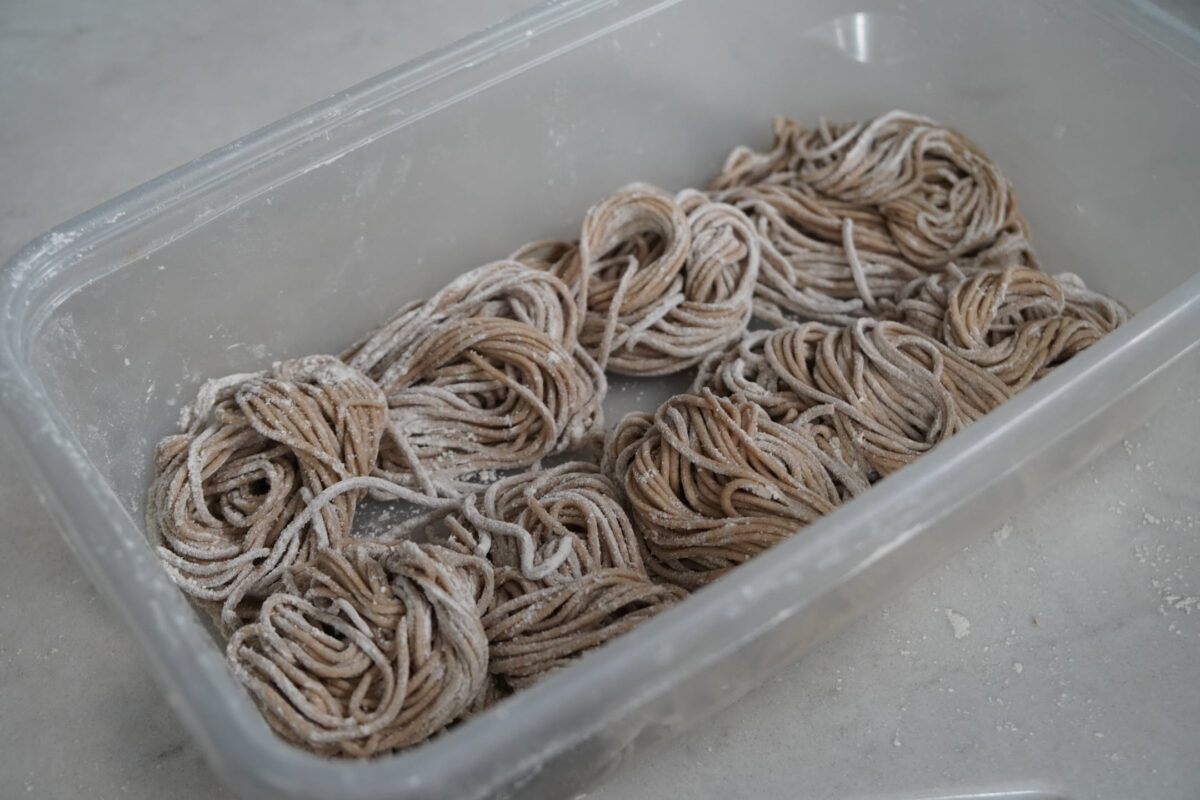 Six coiled nests of fresh handmade soba noodles dusted with flour arranged in a clear plastic storage container on a light countertop