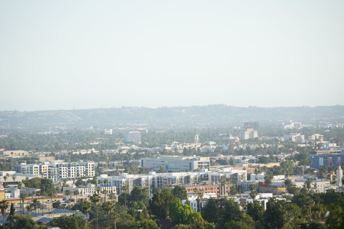 Elevated daytime view over a sprawling urban neighborhood with low-rise buildings, palm trees, and a hazy sky stretching to distant hills