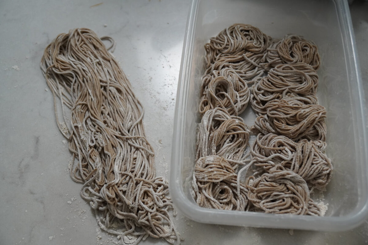 Freshly cut soba noodles dusted with flour, some loosely piled on a floured surface and others coiled into bundles inside a clear plastic container, overhead view on a grey work surface