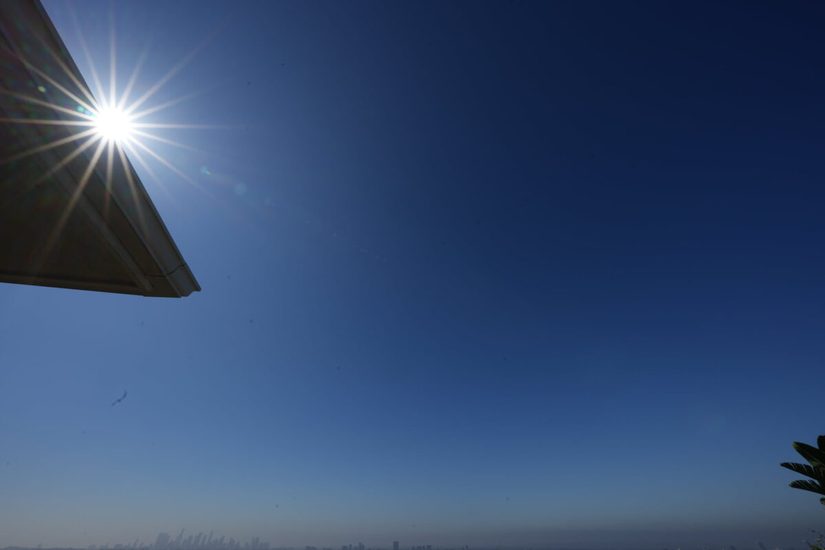 Low-angle shot looking up at a rooftop corner with a bright sun starburst in a clear deep blue sky; distant city skyline visible near the horizon, palm fronds in lower right corner
