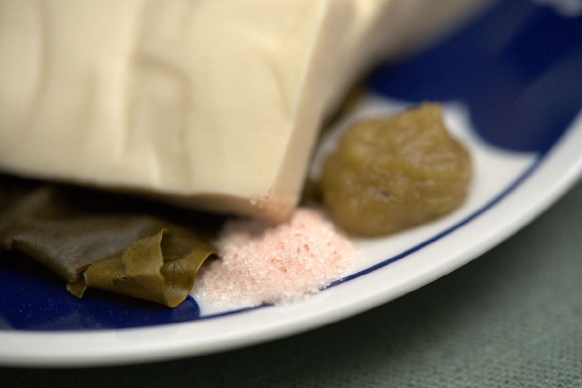 Close-up of a blue-rimmed white plate holding a block of firm tofu, dark green kombu seaweed, pink himalayan salt, and a small piece of pickled or preserved vegetable