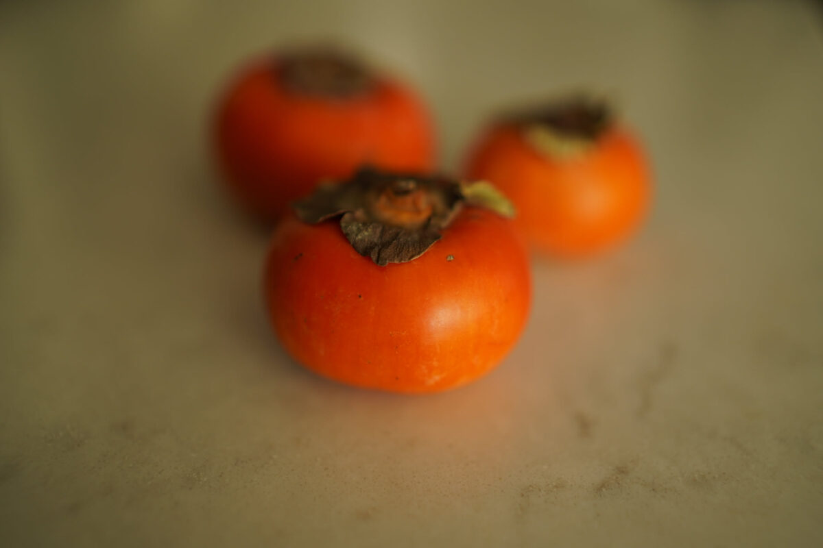 Three vibrant orange persimmons with dried brown calyx leaves arranged in a loose cluster on a neutral gray surface, foreground fruit in sharp focus with shallow depth of field blurring the two behind it