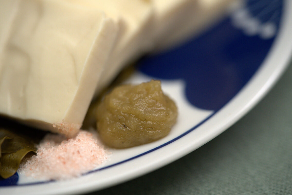 Close-up of a blue-rimmed white plate holding sliced firm tofu, a dollop of pale green paste, pink salt crystals, and a piece of kombu seaweed