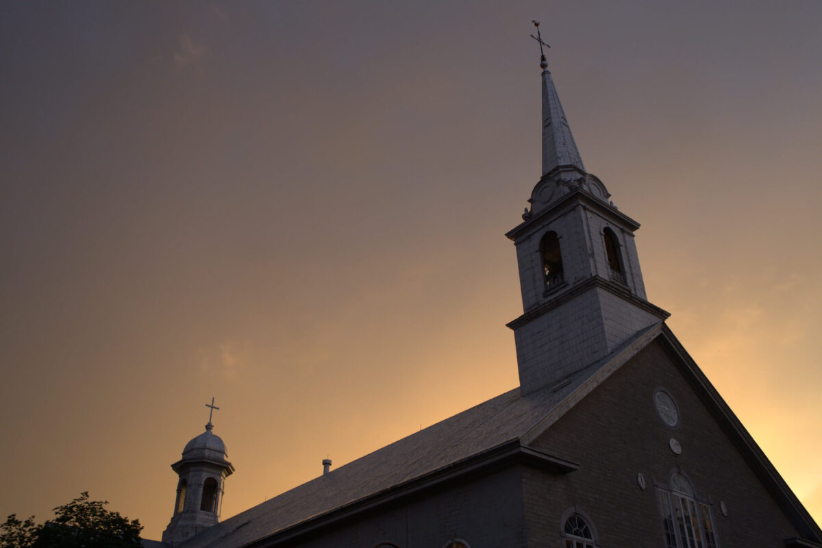 Low-angle view of a stone church with tall white steeple and secondary cupola silhouetted against a warm orange and grey sunset sky