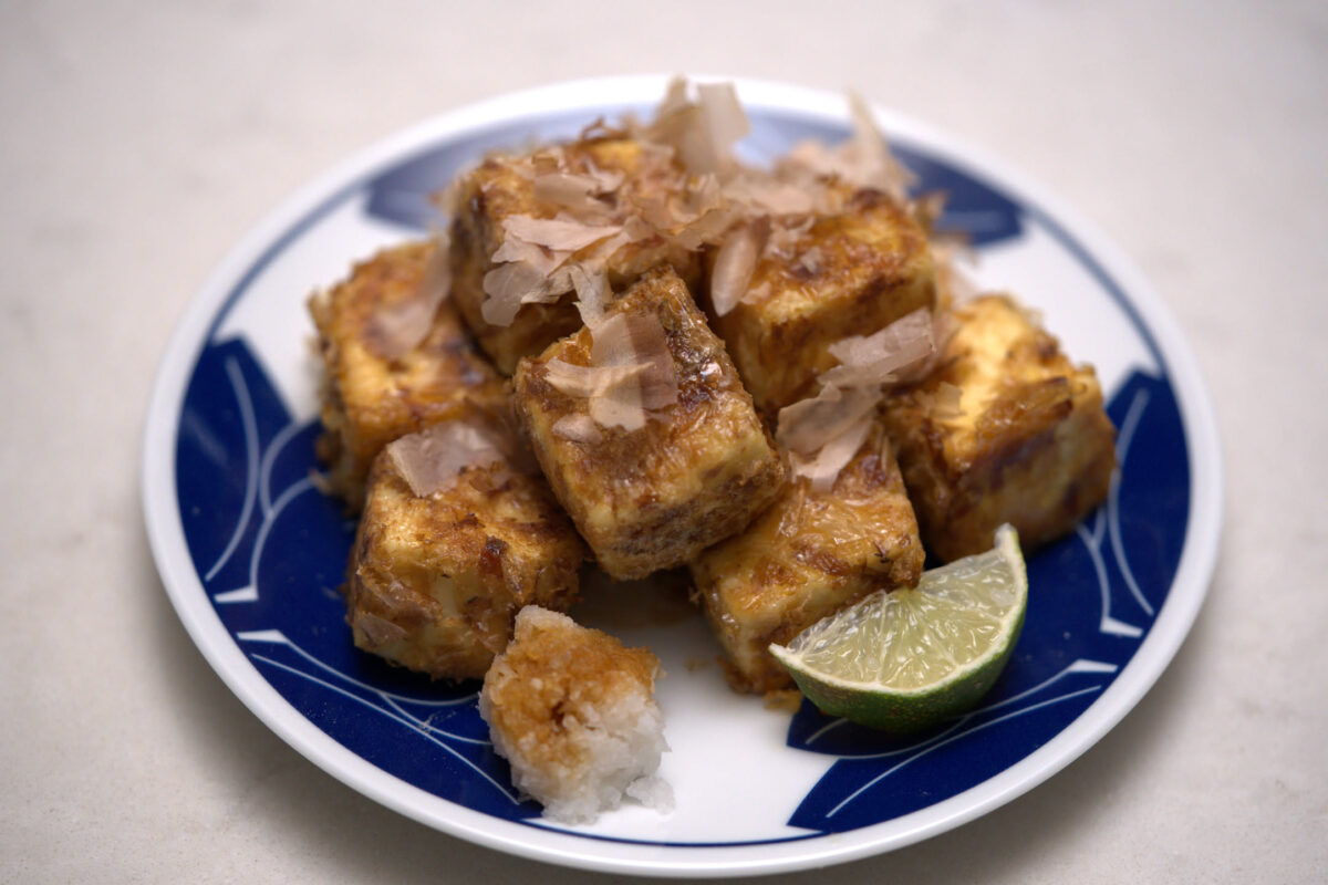 Sliced fried tofu pieces topped with katsuobushi (bonito flakes) served on a blue and white ceramic plate with a lime wedge garnish