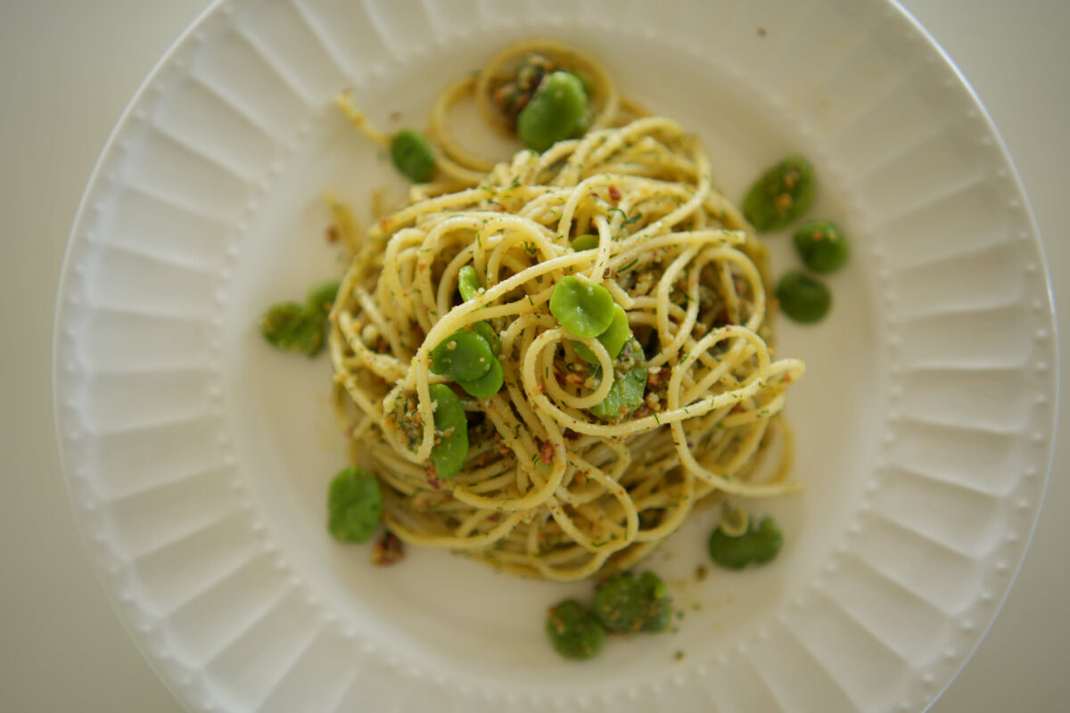 Overhead shot of spaghetti tossed in a green herb and breadcrumb sauce, garnished with shelled fava beans, plated on a white ridged plate