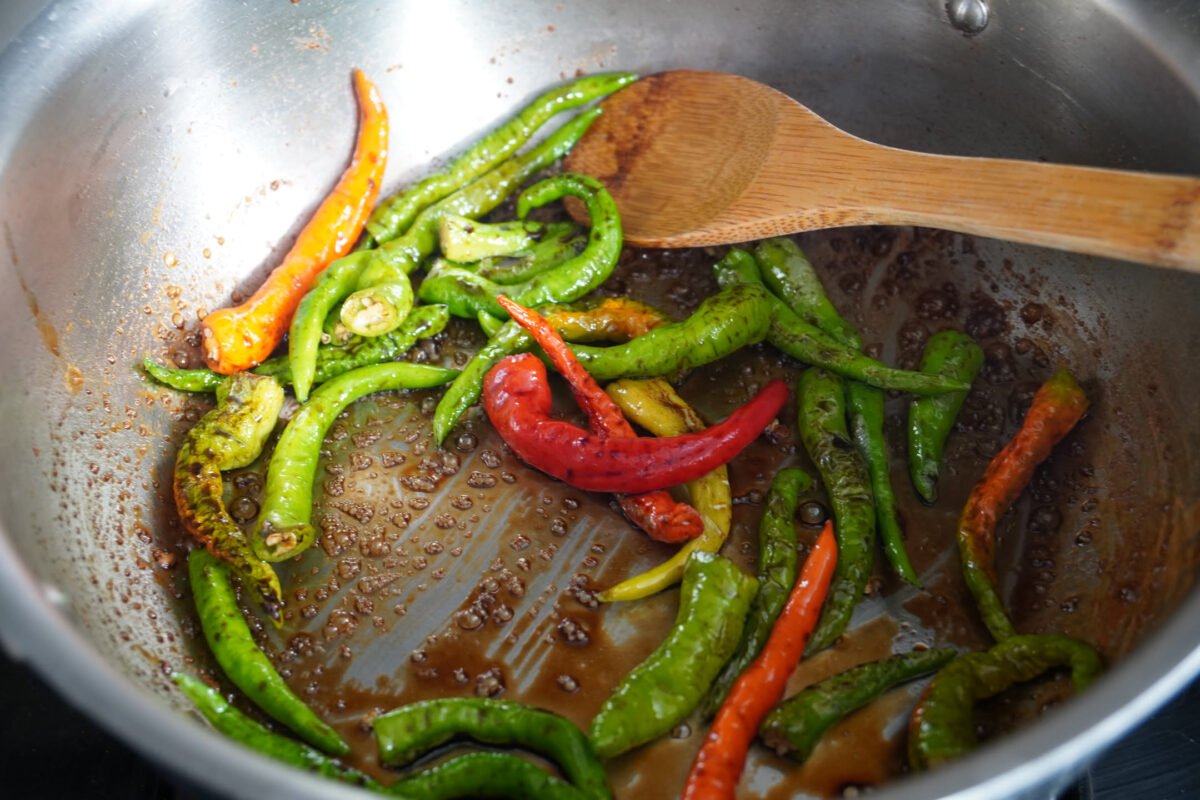 Green and red chili peppers sautéing in a stainless steel pan with dark sauce, wooden spatula resting on the rim