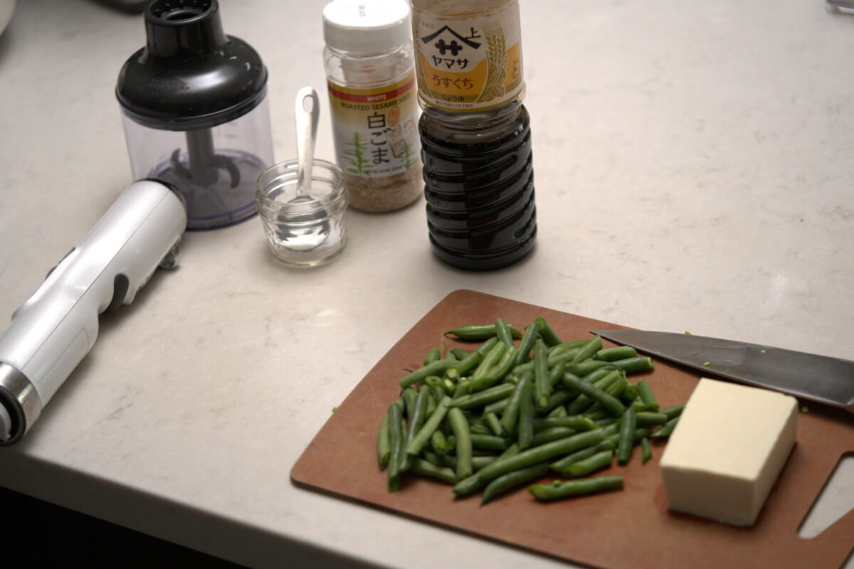 Brown wooden cutting board on a kitchen counter with chopped green beans, a block of tofu, and a chef's knife, alongside Japanese condiments including Yamasa soy sauce and sesame seeds