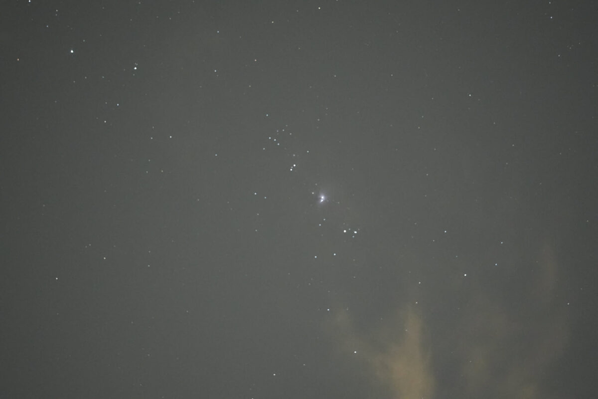Night sky showing Orion constellation with bright Orion Nebula visible at center, scattered stars against dark grey sky, light glow in lower right corner