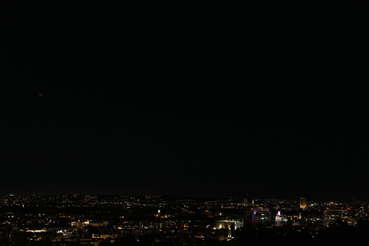 Wide elevated nighttime view of a sprawling city with scattered building lights and illuminated structures against a dark sky