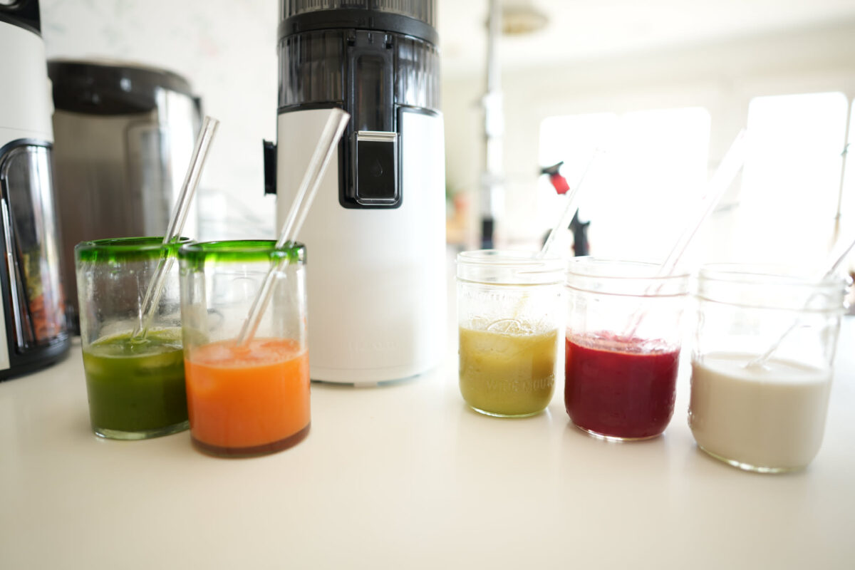 Five glasses and mason jars of freshly pressed juices in green, orange, yellow-green, deep red, and white arranged on a white kitchen counter beside a cold-press juicer, with glass straws in each