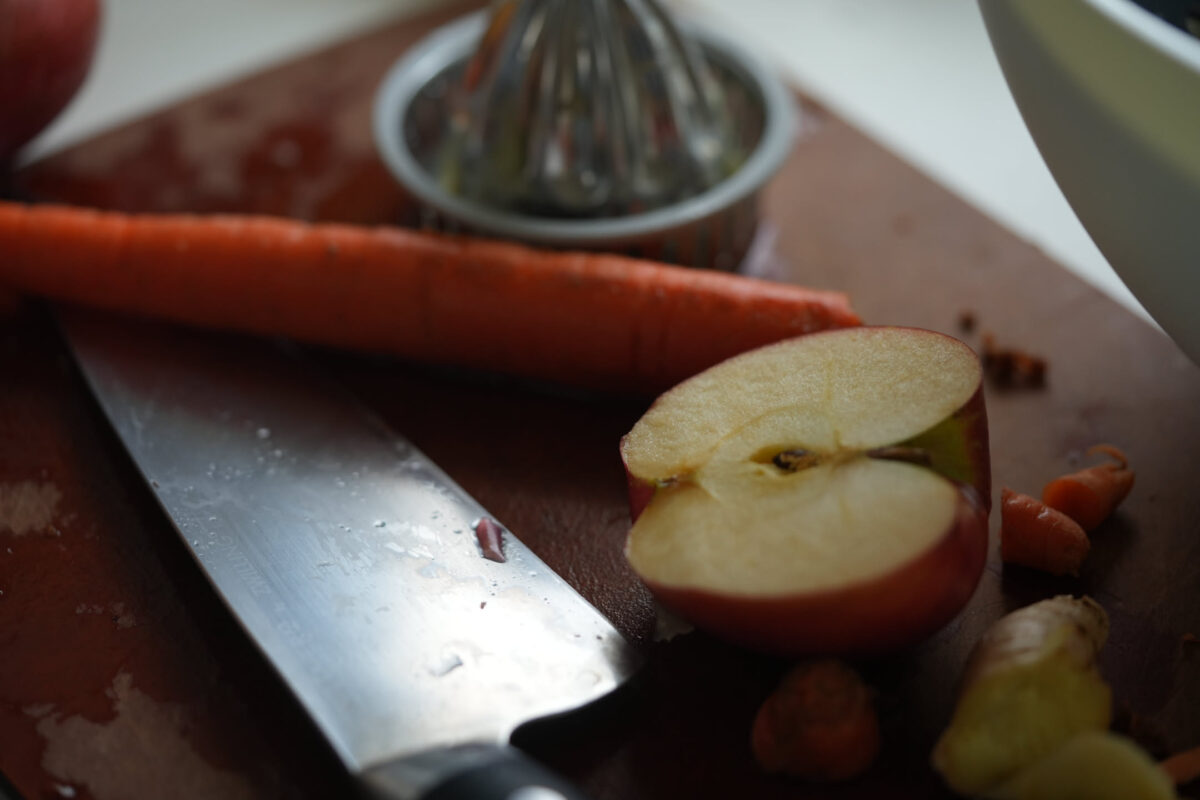 Close-up of a chef's knife blade resting on a dark wooden cutting board beside a halved apple, a whole carrot, small nuts, and a metal citrus juicer in the background