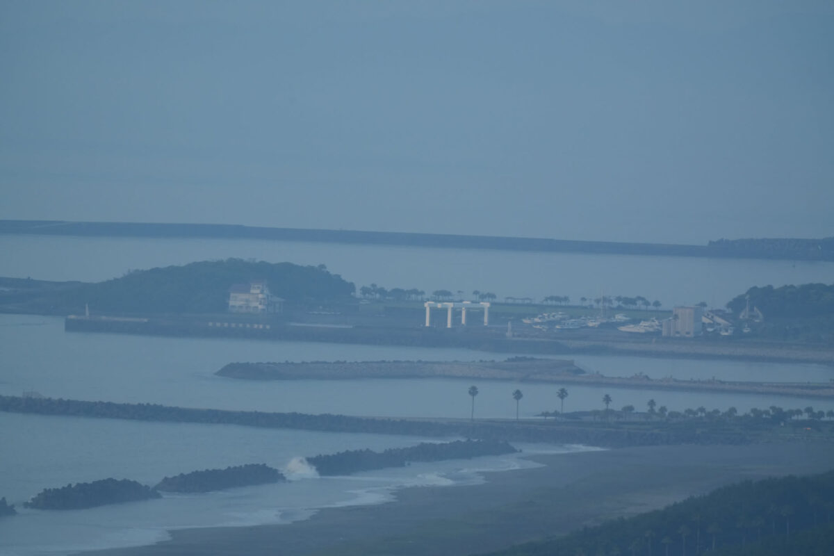 Aerial telephoto view of a hazy coastal bay showing layered sandbars, breakwaters, palm trees, and a small harbor with white columned structure, shot in flat blue-grey light