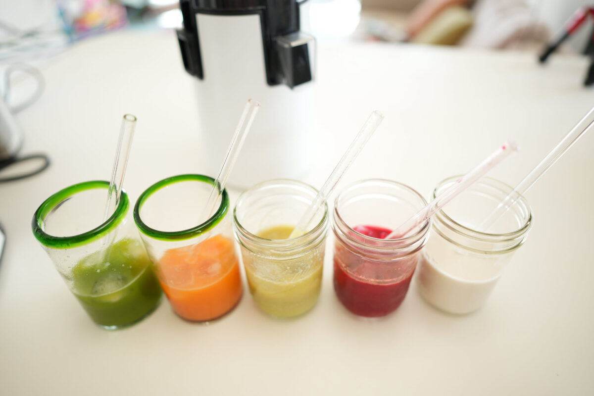 Five small glasses and mason jars in a row on a white table, each containing a different freshly made juice — green, orange, pale yellow, deep red, and white — with glass straws, a juicer in the background