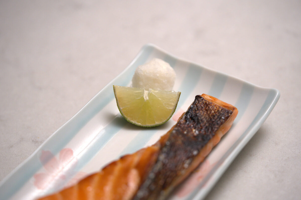 Grilled salmon fillet on a blue-striped Japanese ceramic plate, accompanied by a wedge of small green citrus (sudachi or kabosu) and a mound of grated daikon radish
