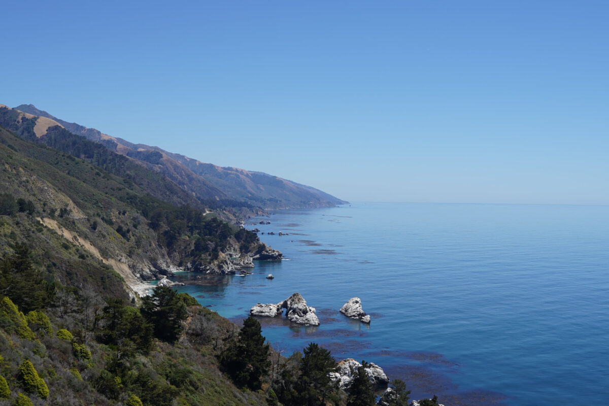 Aerial view of rugged California coastline with white sea stacks, turquoise coves, tree-covered hillsides, and mountain ridgeline stretching into the distance under clear blue sky