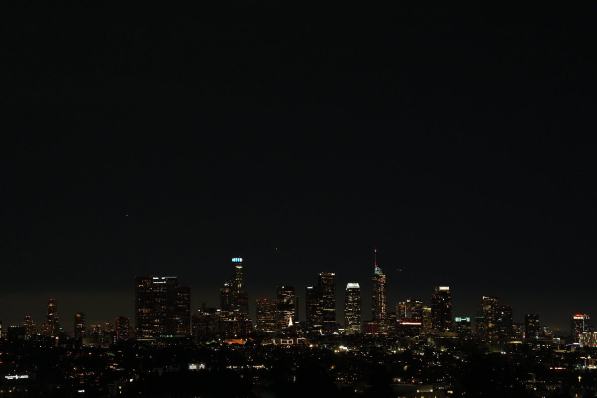 Downtown Los Angeles skyline at night with illuminated skyscrapers against a dark sky, shot from an elevated vantage point with foreground rooftops in silhouette