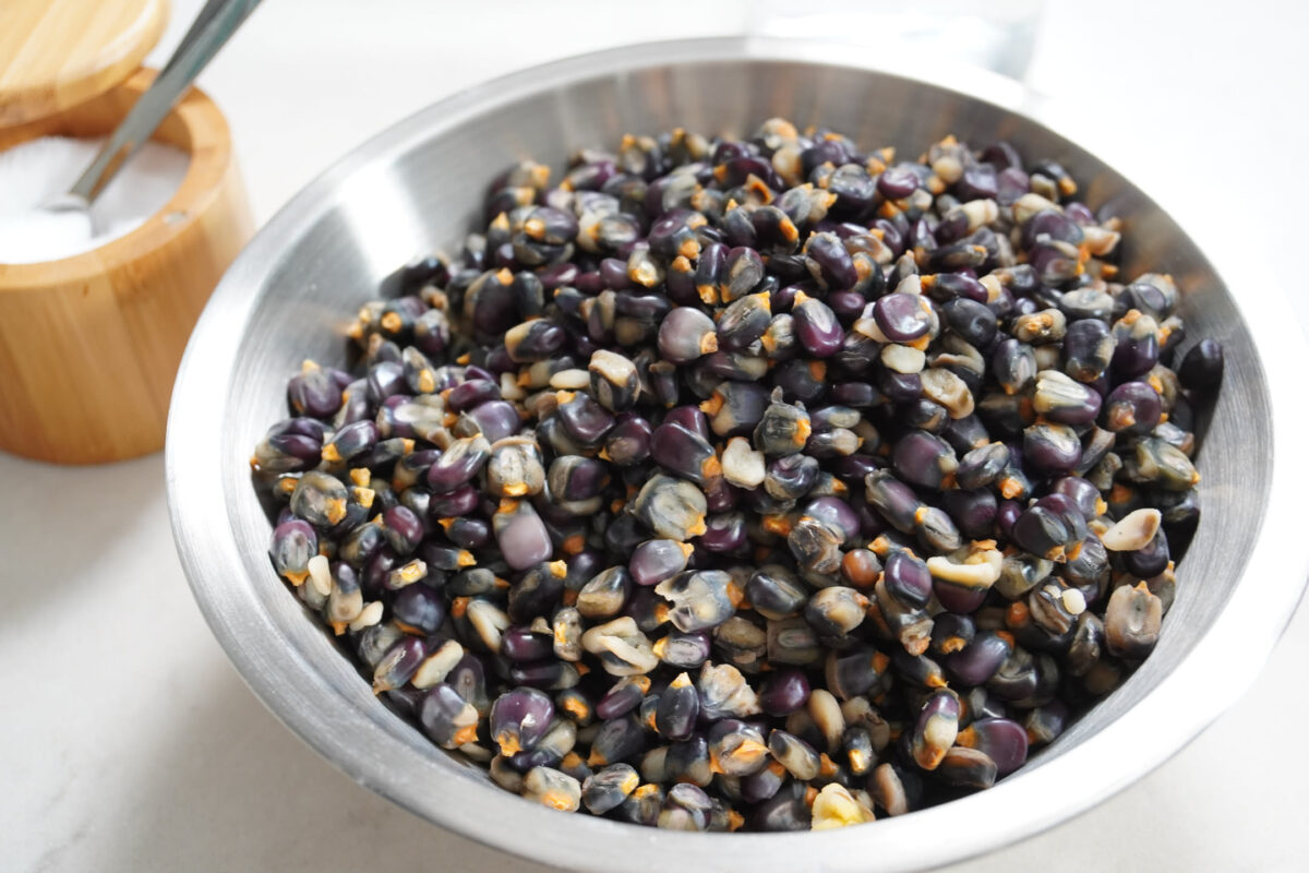 Stainless steel mixing bowl filled with raw purple and black corn kernels, with a wooden salt box and spoon in the background on a white surface