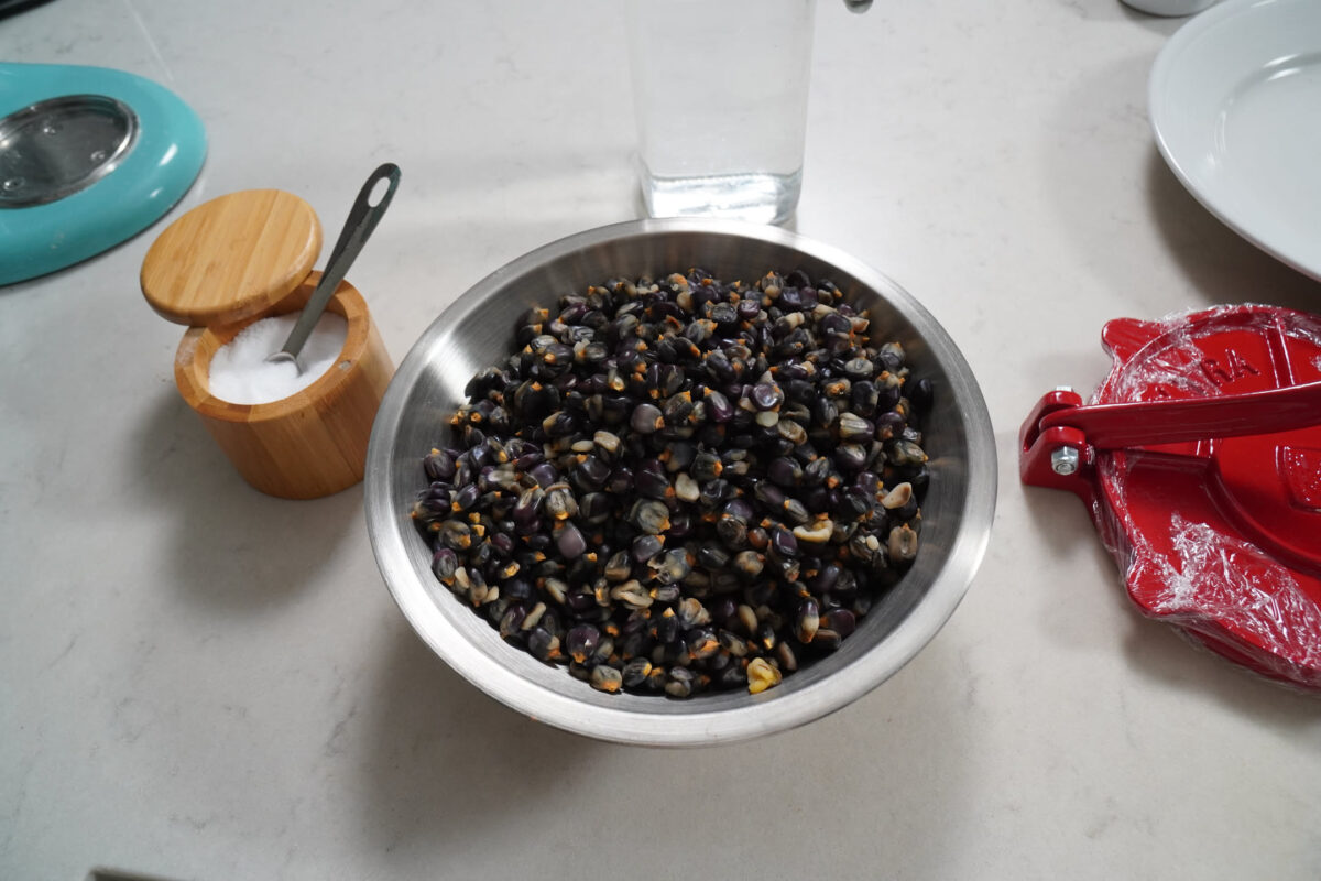 Stainless steel mixing bowl filled with dark purple-black corn kernels on a white marble countertop, with a wooden salt cellar, red corn stripper tool, and glass in the background