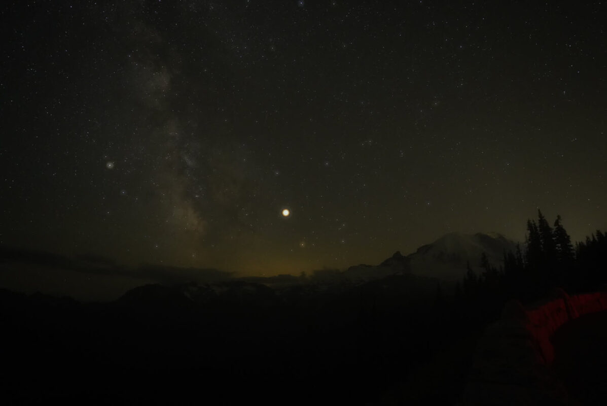 Night sky showing the Milky Way galaxy and a bright planet or moon above a dark mountain and conifer tree silhouette with a warm yellowish glow near the horizon