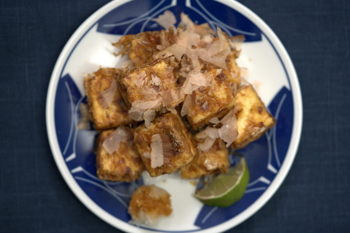 Overhead shot of golden fried tofu cubes topped with bonito flakes served on a blue-and-white Japanese plate with a small lime wedge on the side