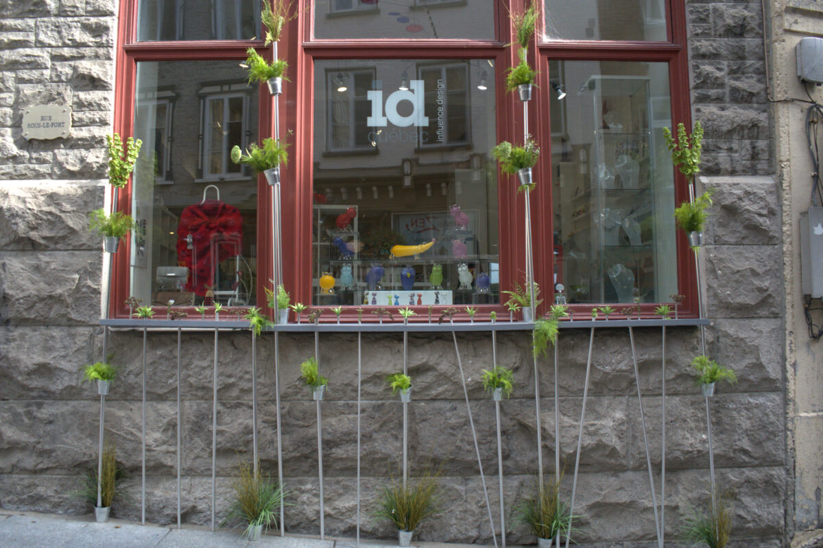 Storefront of a shop with a red window frame, displaying a metal fence/railing along the base with small potted plants and climbing greenery, stone wall facade, urban street setting