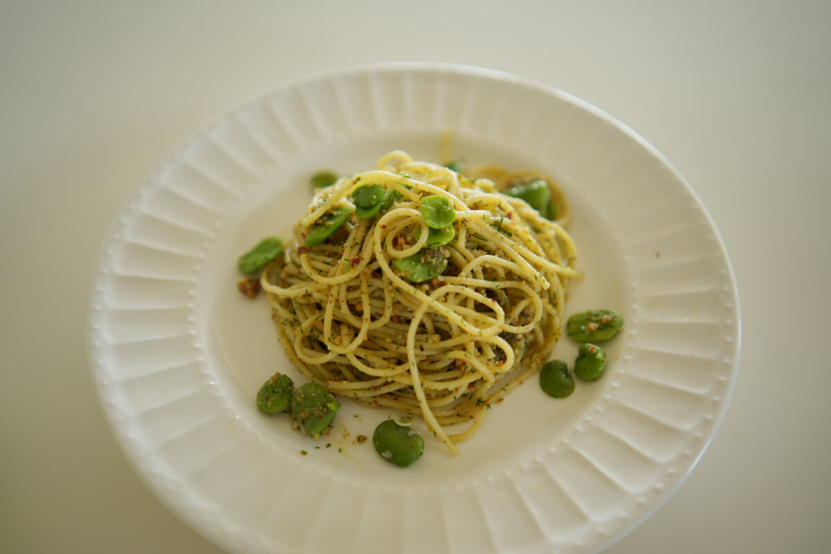 Overhead view of spaghetti tossed in a green herb sauce with shelled fava beans scattered around, served on a white ridged plate against a light background